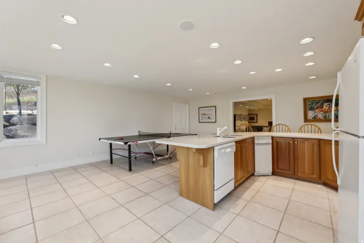 Recessed lighting throughout, tile flooring, kitchen island with integrated sink, wood cabinetry, and a pass-through window to an additional space