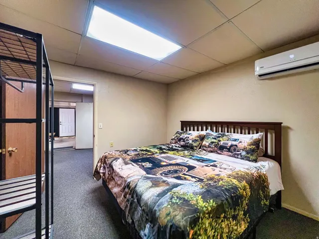 Recessed lighting, drop ceiling, wall-mounted air conditioning unit, wood-finish bed frame, and carpeted flooring