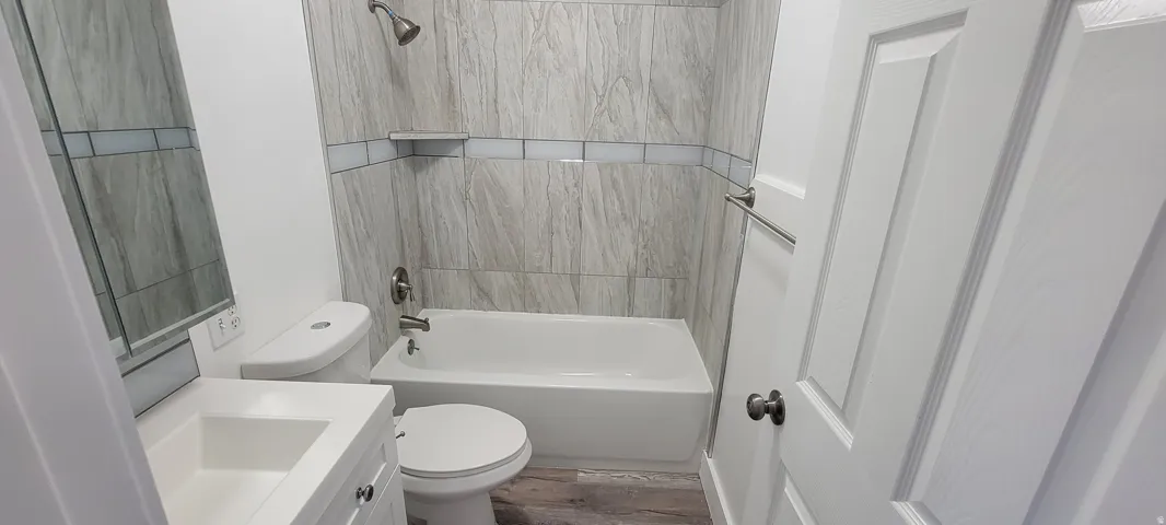 White vanity with integrated sink, wood-finish flooring, tiled shower surround with accent band, and a shower/tub combination