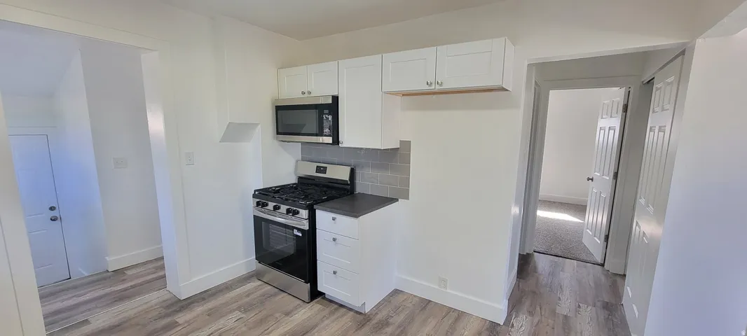 Kitchen area featuring wood-finish flooring, white cabinetry, stainless steel microwave, gas range, and gray subway tile backsplash