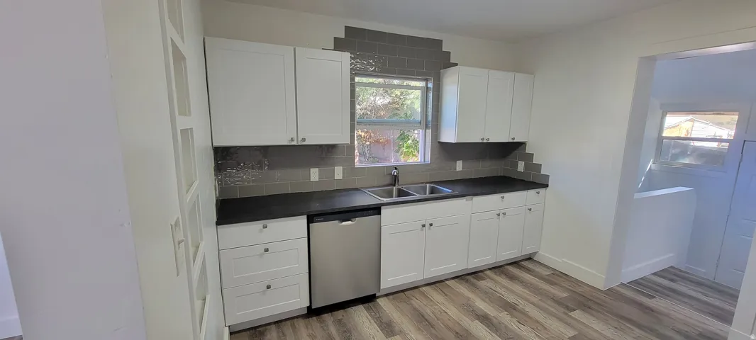White shaker cabinetry, stainless steel dishwasher, dark countertops, wood-finish flooring, and a tiled backsplash