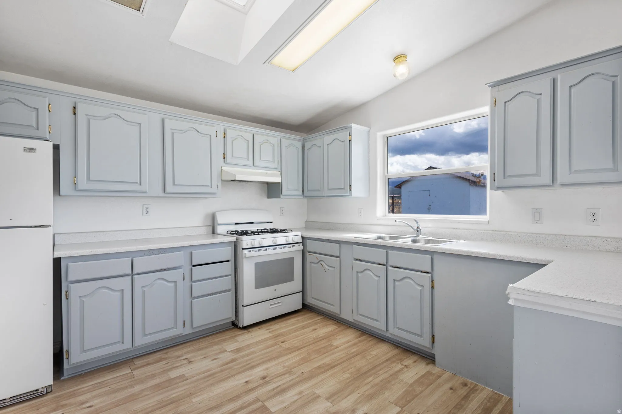 Light blue cabinetry with white countertops, white appliances, wood-finish flooring, and a skylight