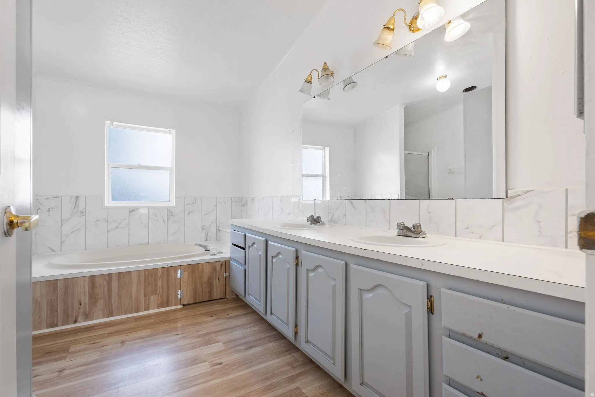 Dual vanity with white countertops, wood-finish flooring, a large soaking tub, and a window for natural light