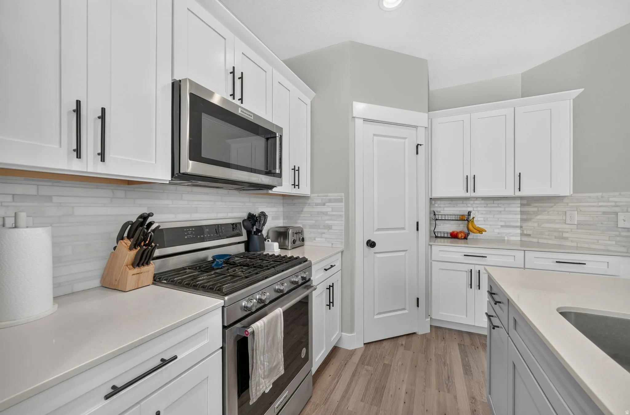 White cabinetry with matte black hardware, stainless steel appliances, light-toned countertops, tile backsplash, and wood-finish flooring