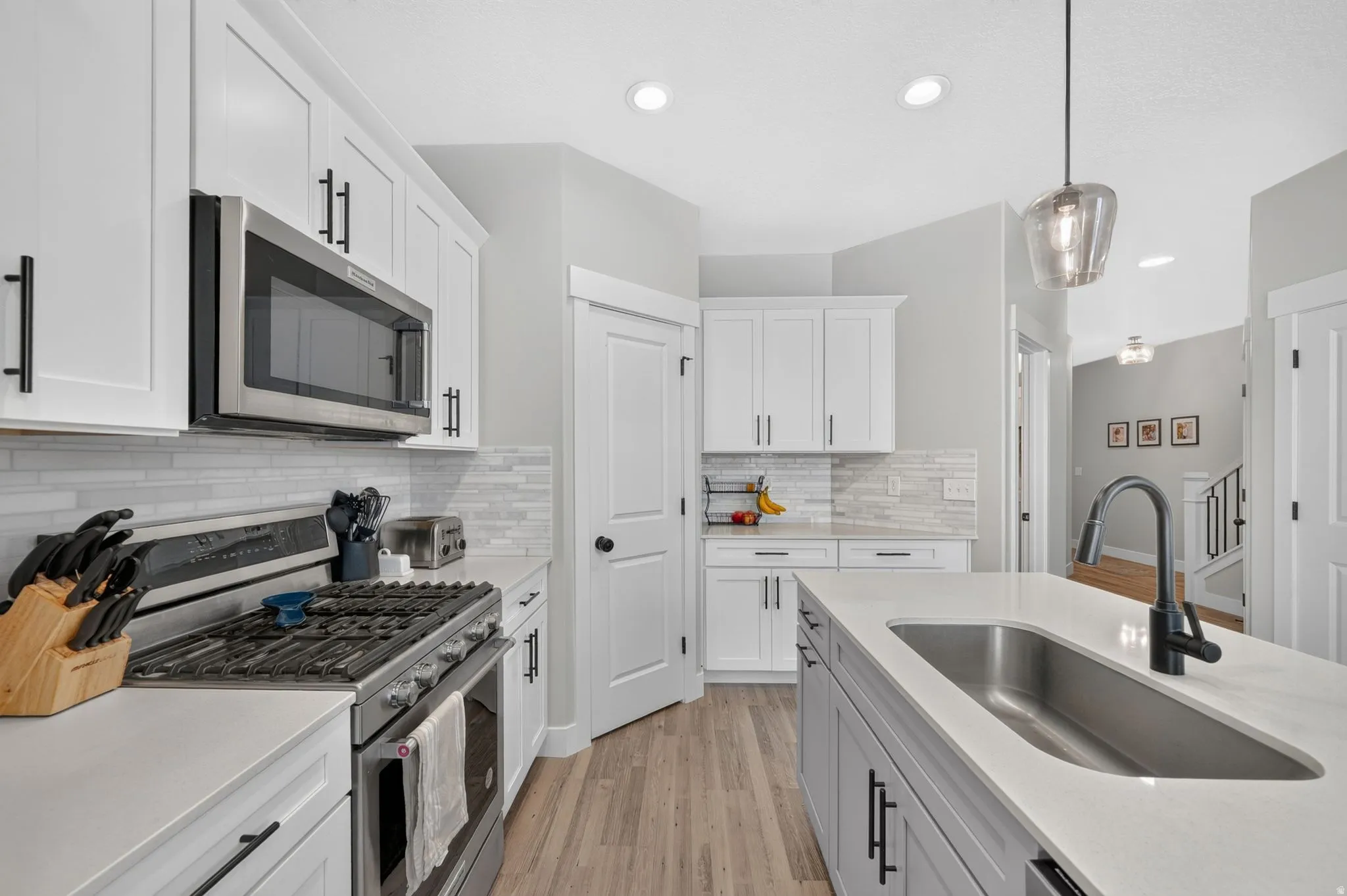 Stainless steel gas range with microwave, white shaker cabinetry, light-toned wood-finish flooring, island with undermount sink, and recessed lighting