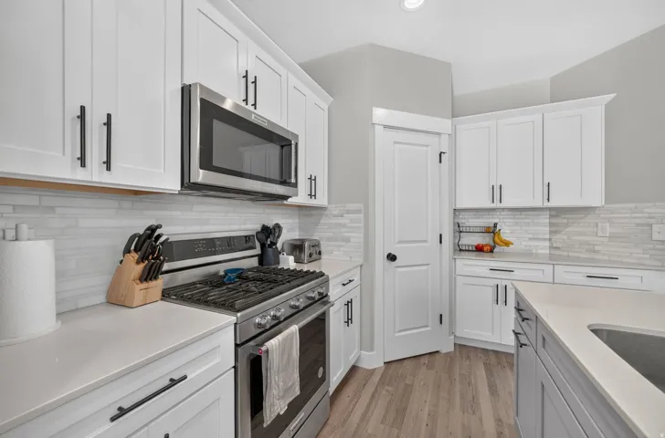 White cabinetry with matte black hardware, stainless steel appliances, light-toned countertops, tile backsplash, and wood-finish flooring