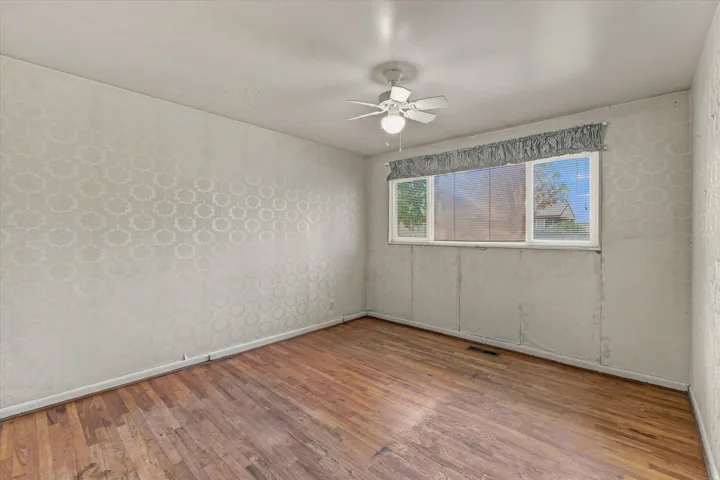 Wood-finish flooring, patterned wallpaper, a ceiling fan with integrated lighting, and a single window with a valance