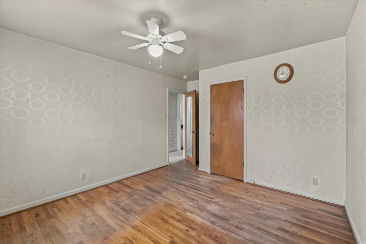 Wood-finish flooring, patterned wallpaper, ceiling fan with integrated lighting, and a solid wood door