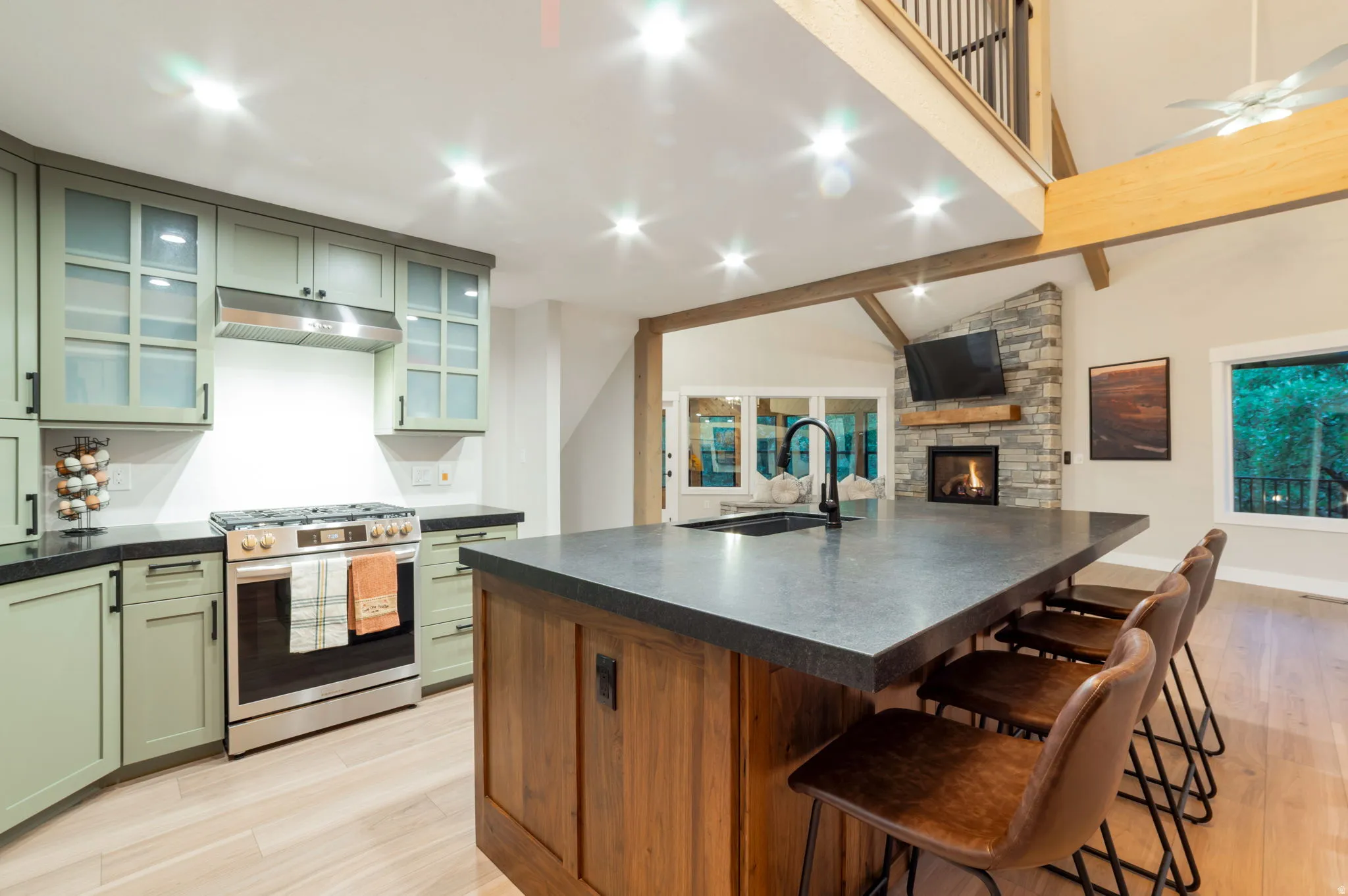 Kitchen featuring light green cabinetry, stainless steel range and hood, a substantial island with a dark countertop and integrated sink, and light wood-finish flooring