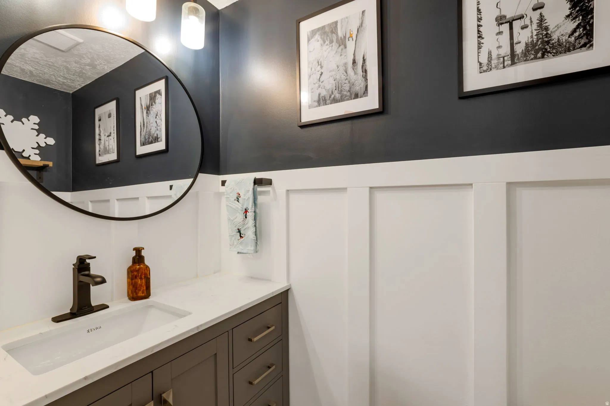Board and batten wainscoting, a single vanity with a white countertop, an integrated rectangular sink, a dark bronze faucet, and a circular mirror