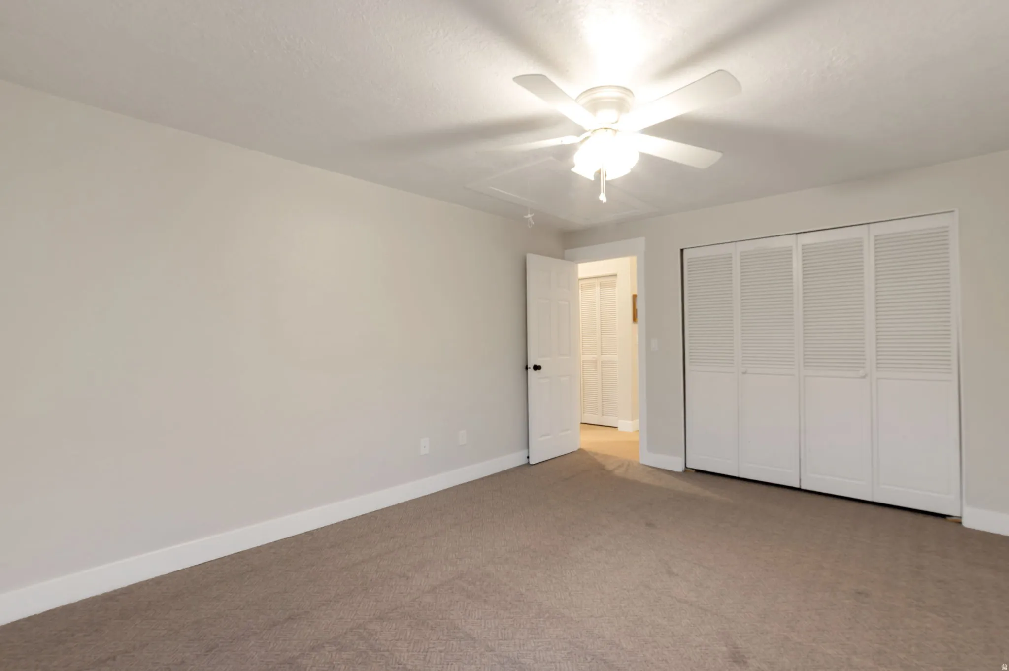Spacious bedroom with neutral wall finishes, neutral-toned carpet flooring, ceiling-mounted light fixture with fan, and louvered closet doors providing ample storage