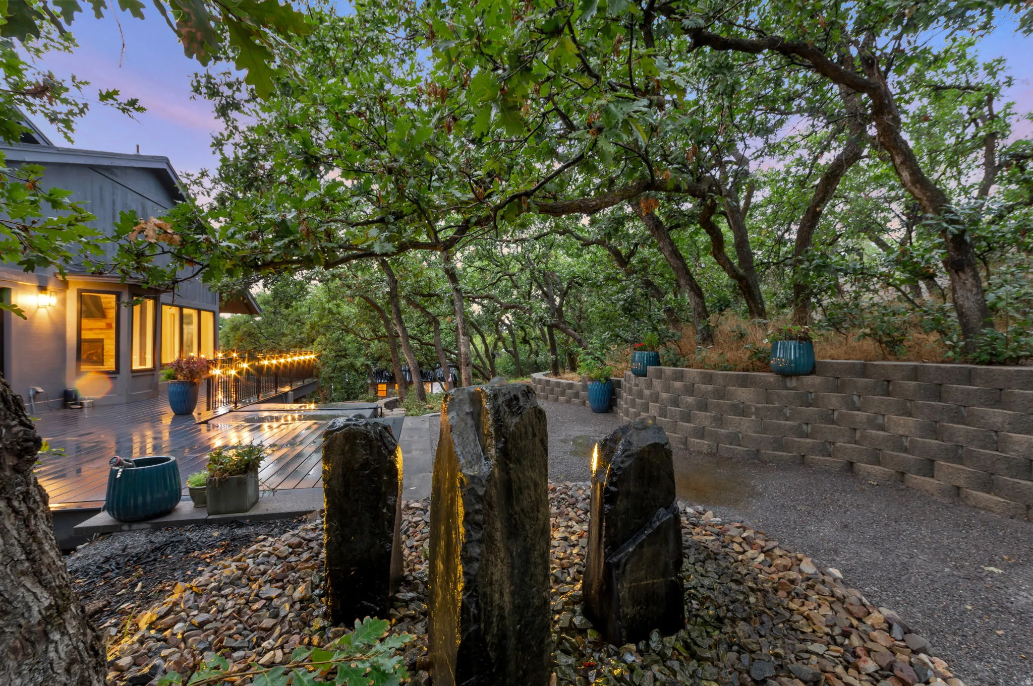 Expansive outdoor decking, mature trees providing natural canopy, retaining wall, natural stone fountain, and a gravel pathway.