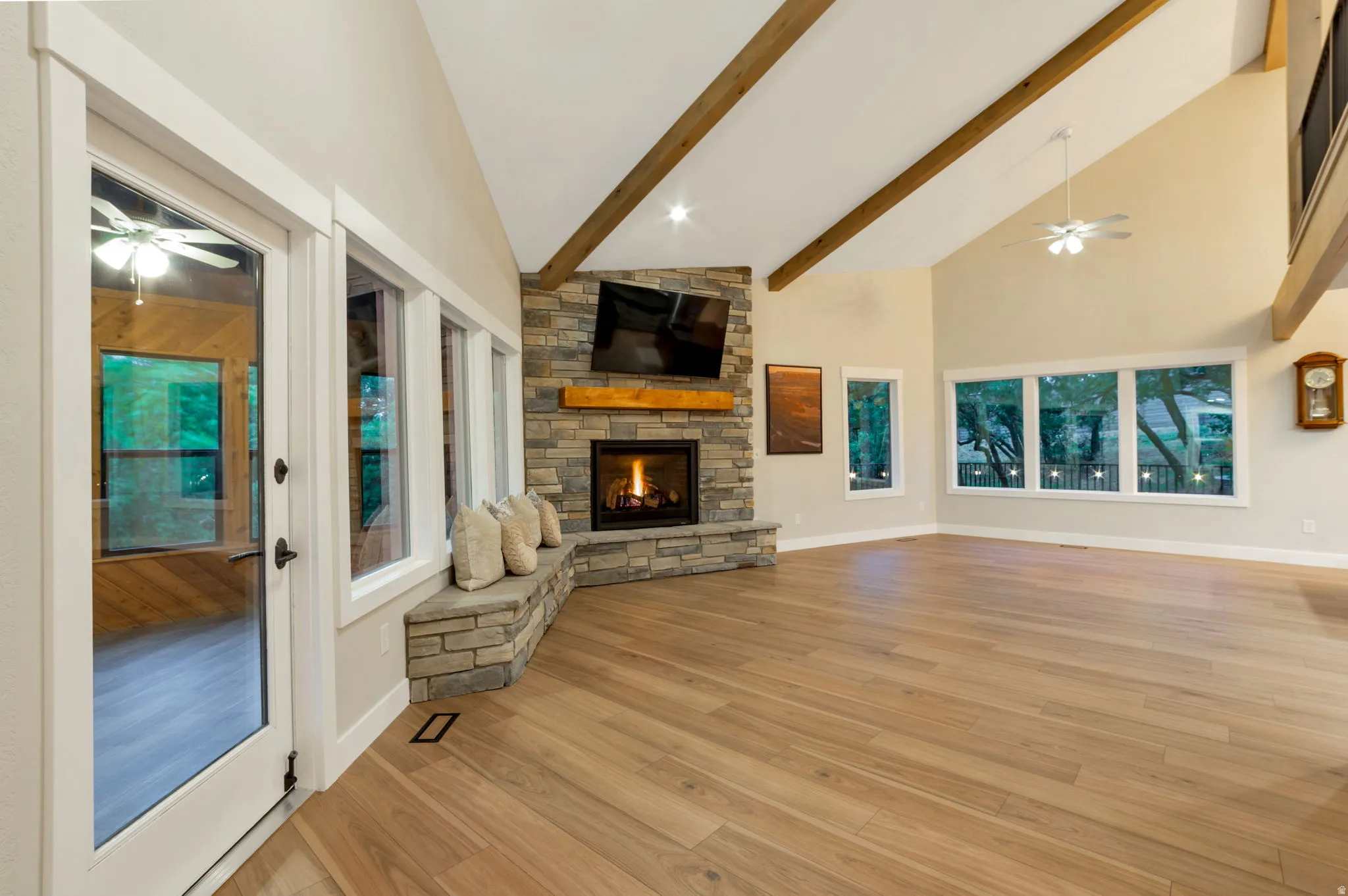Vaulted ceiling with exposed wood beams, stone fireplace with a wood mantle, stone bench seating, and multiple picture windows