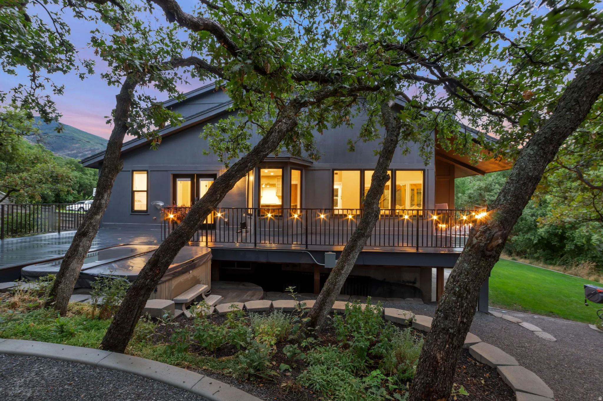 Exterior featuring a gray facade, elevated deck with dark railing, a bay window, and surrounding mature trees.