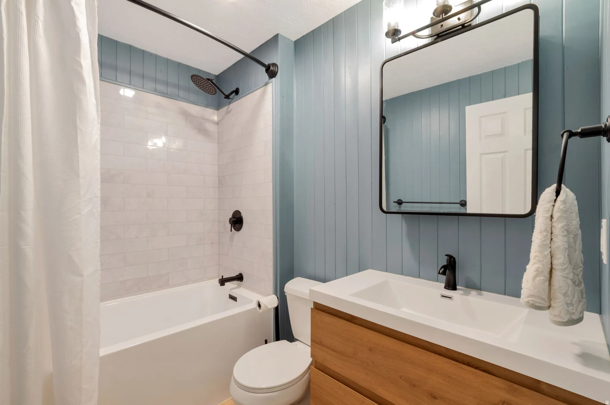 Newley renovated bathroom featuring vertical paneling, matte black fixtures, an integrated sink, a wood-finish vanity, and tub shower.