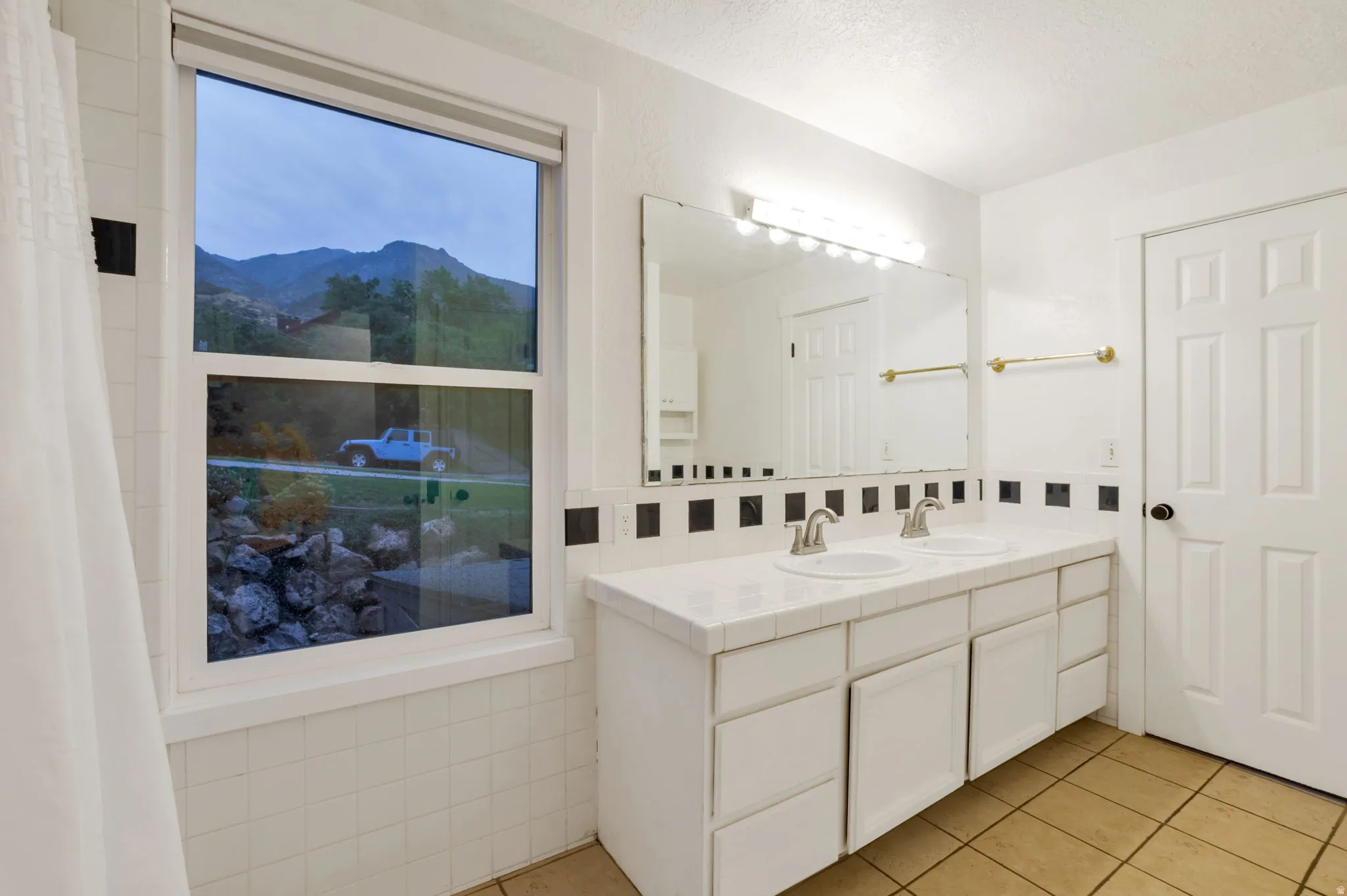 Dual vanity with integrated sinks, large wall-mounted mirror, natural light, white paneled cabinetry, and tiled flooring