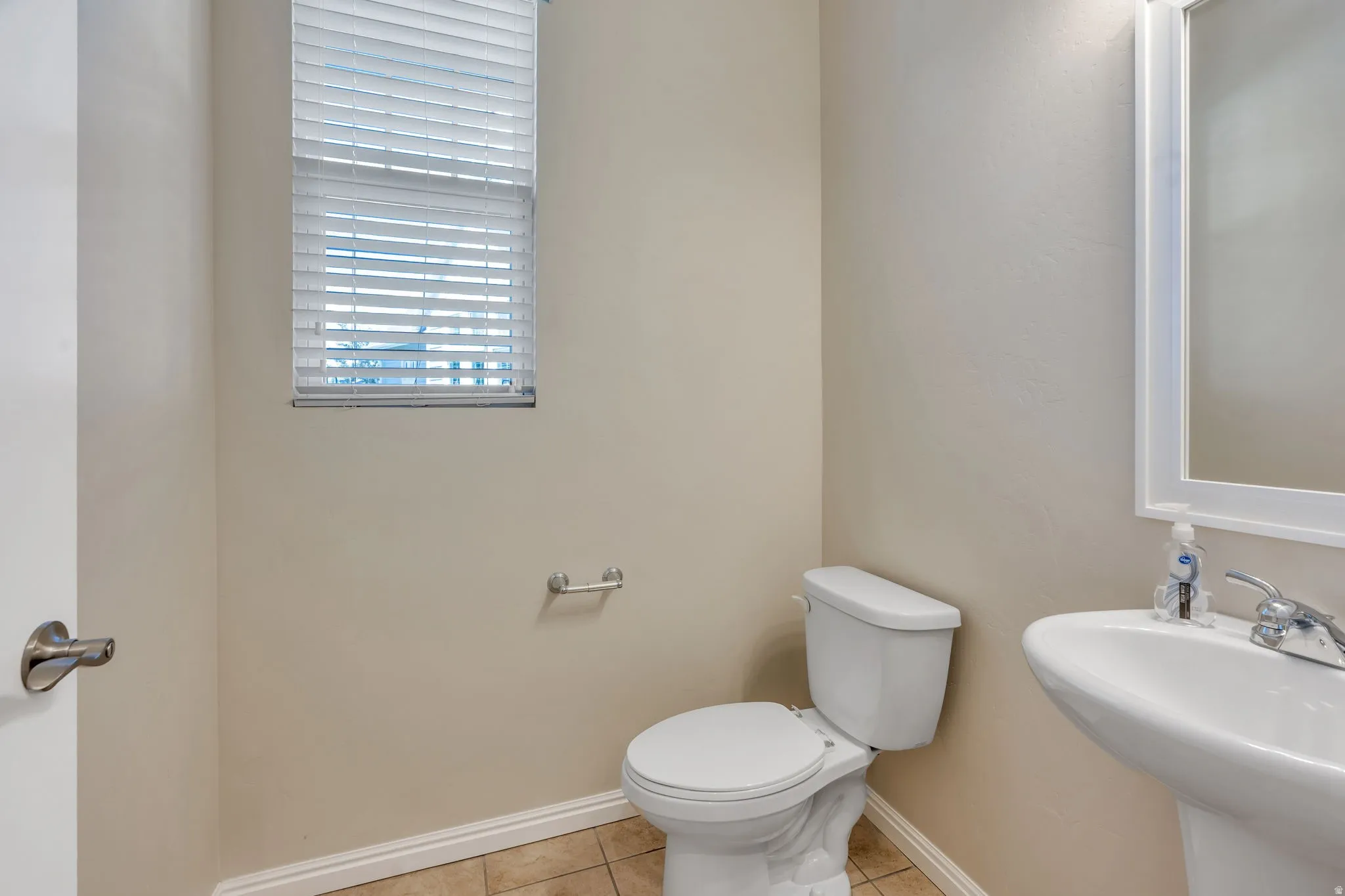 Pedestal sink with chrome faucet, wall-mounted mirror, ceramic tile flooring, and a window with blinds