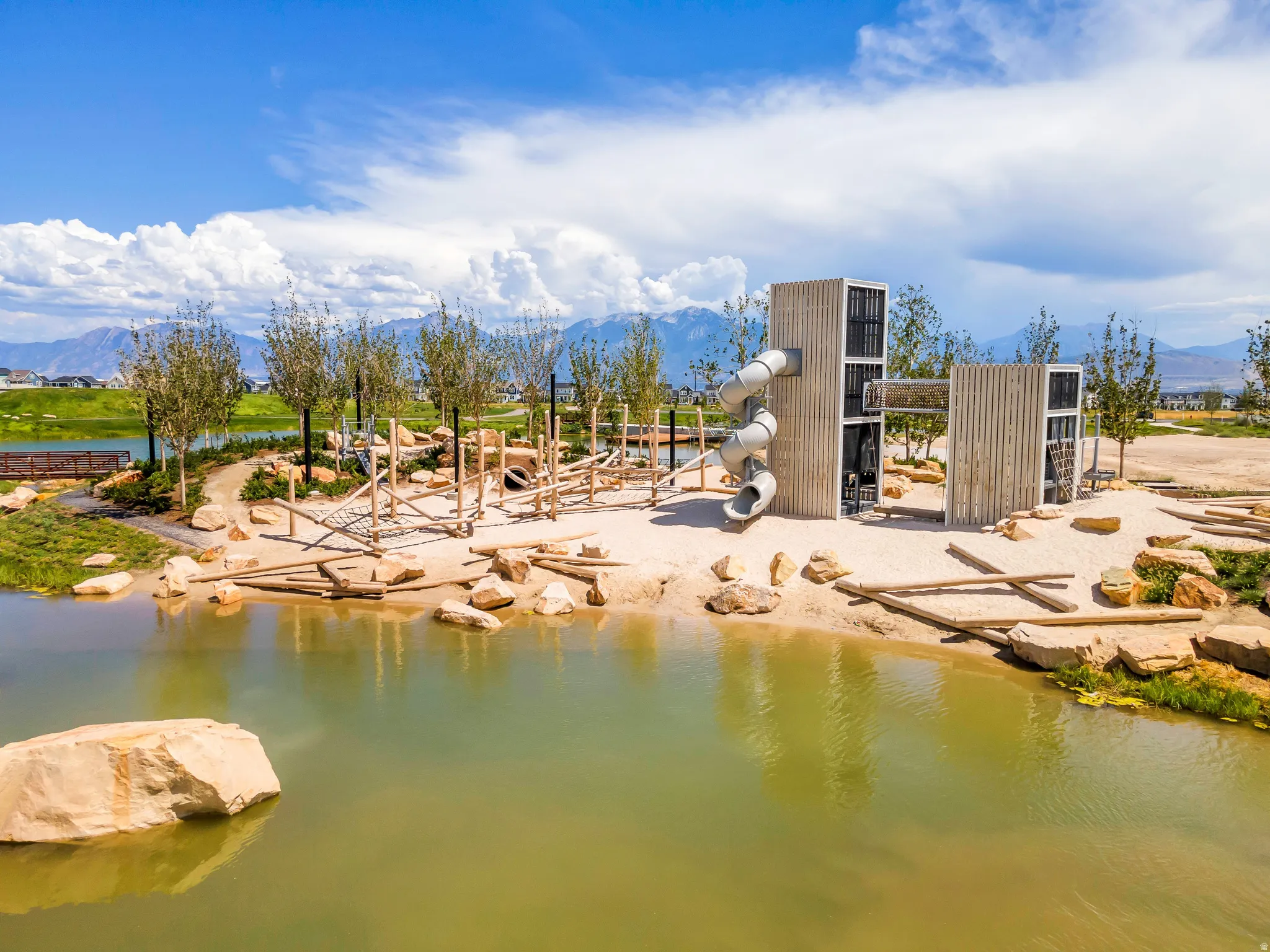 Expansive outdoor recreation area featuring a natural-themed playground with slides and climbing structures, surrounded by a pond with rock accents