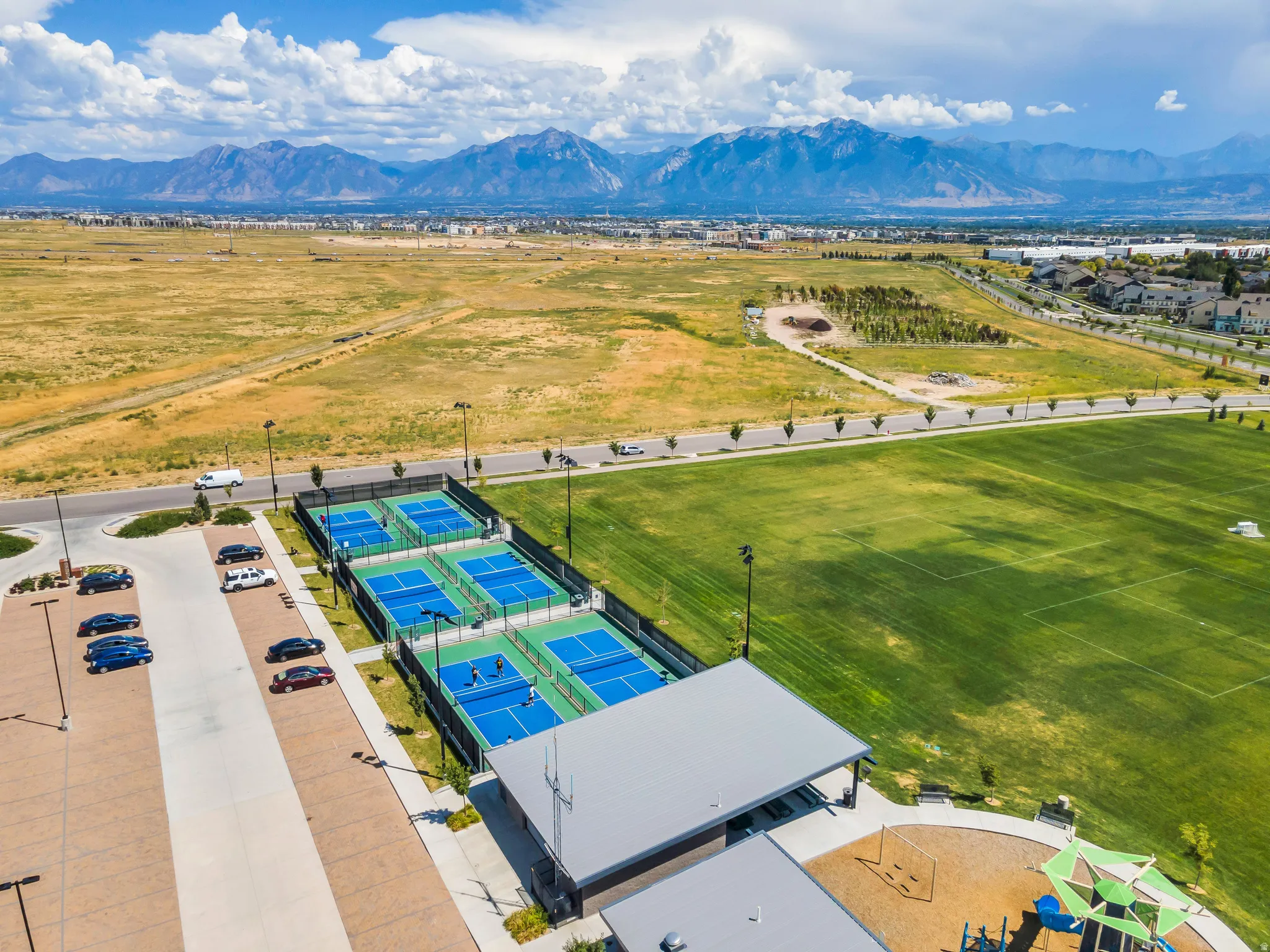 Multiple outdoor pickleball courts, playground with shade structures, expansive green sports fields, and mountain scenery