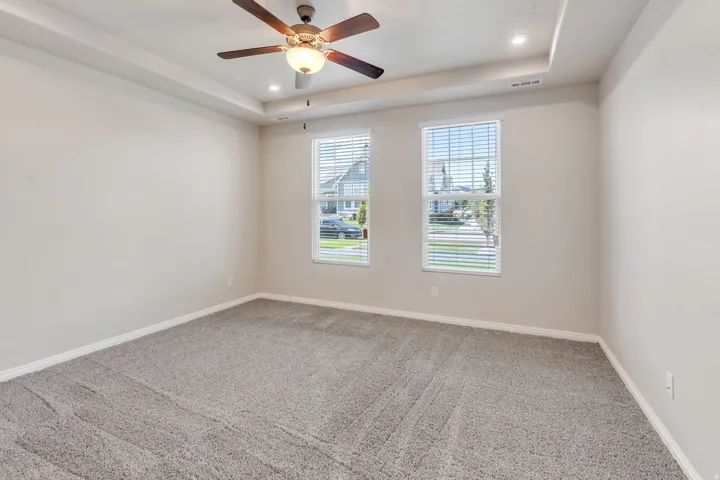 Tray ceiling with recessed lighting, ceiling fan with integrated light, two windows with blinds, and gray textured carpeting