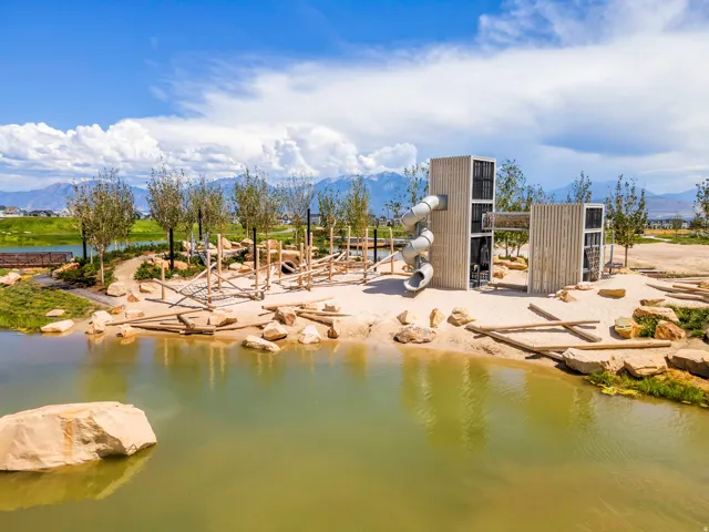 Expansive outdoor recreation area featuring a natural-themed playground with slides and climbing structures, surrounded by a pond with rock accents