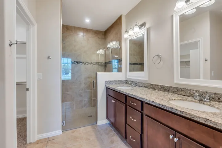 Dual vanity with granite countertops, wood-finish cabinetry, a walk-in shower with tile surround, and tile flooring