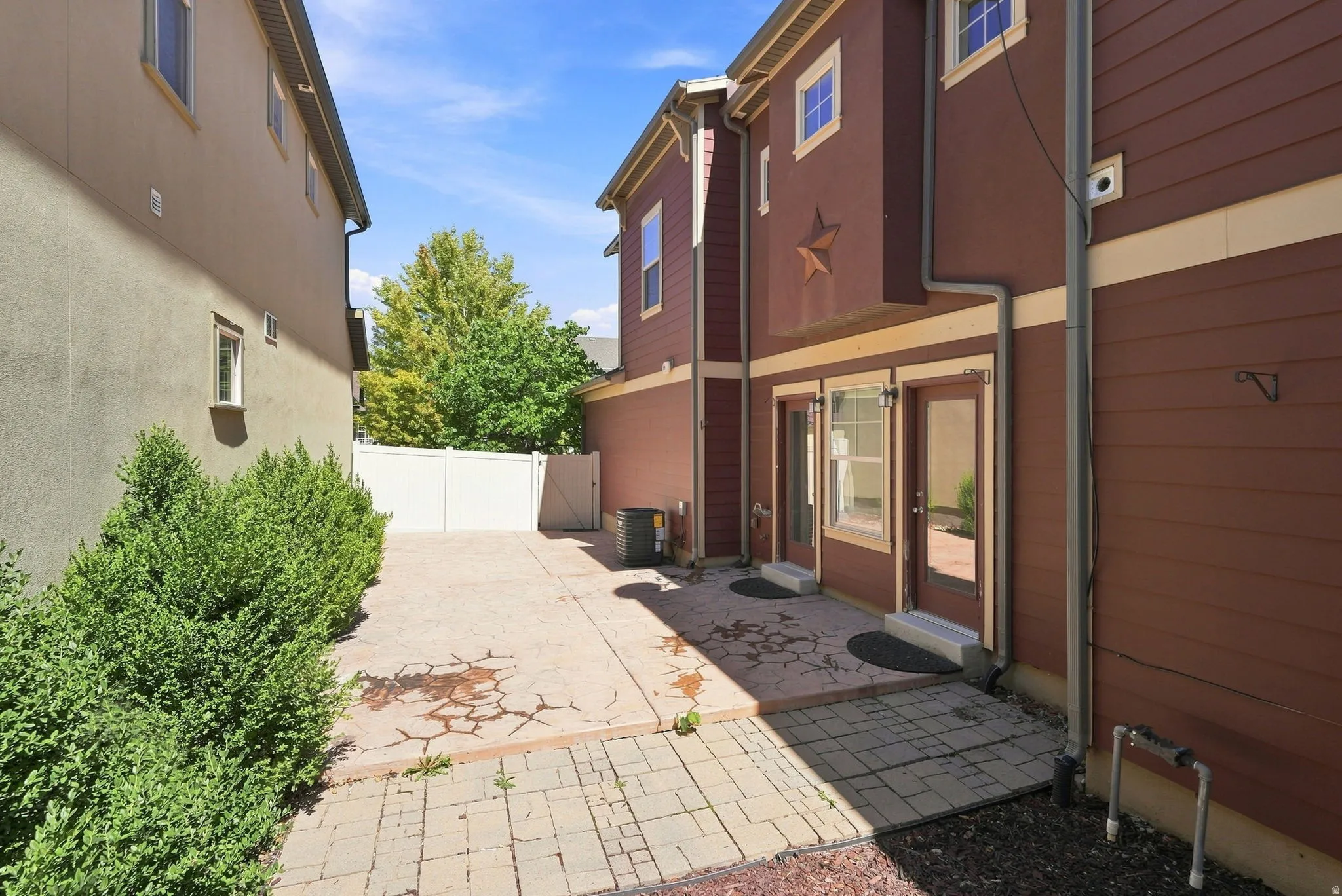 Paved patio area, privacy fence, mature shrubbery, dual glass-panel doors, and brick paver walkway