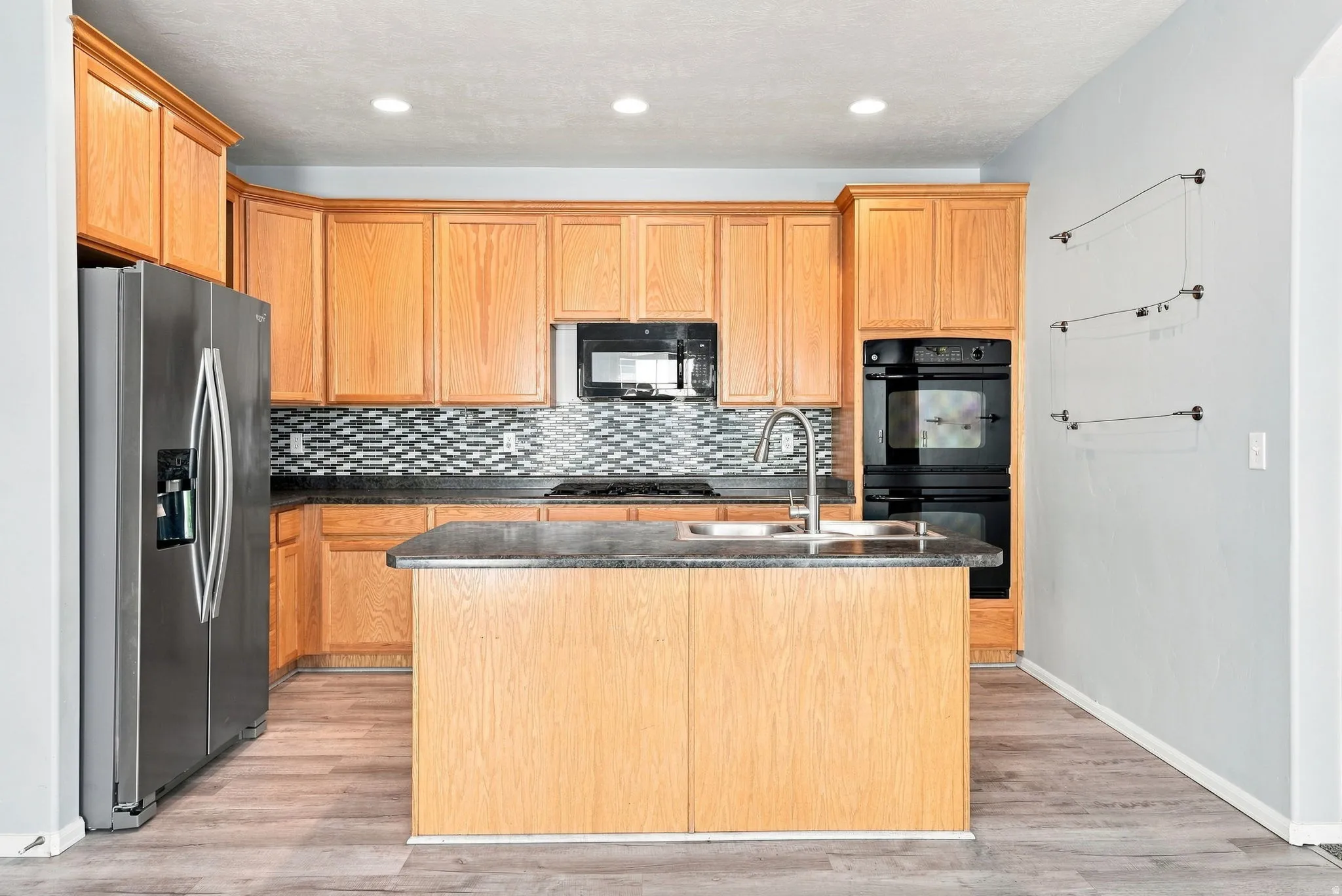 Kitchen featuring wood-finish cabinetry, stainless steel appliances, a center island with a double basin sink, mosaic tile backsplash, and recessed lighting