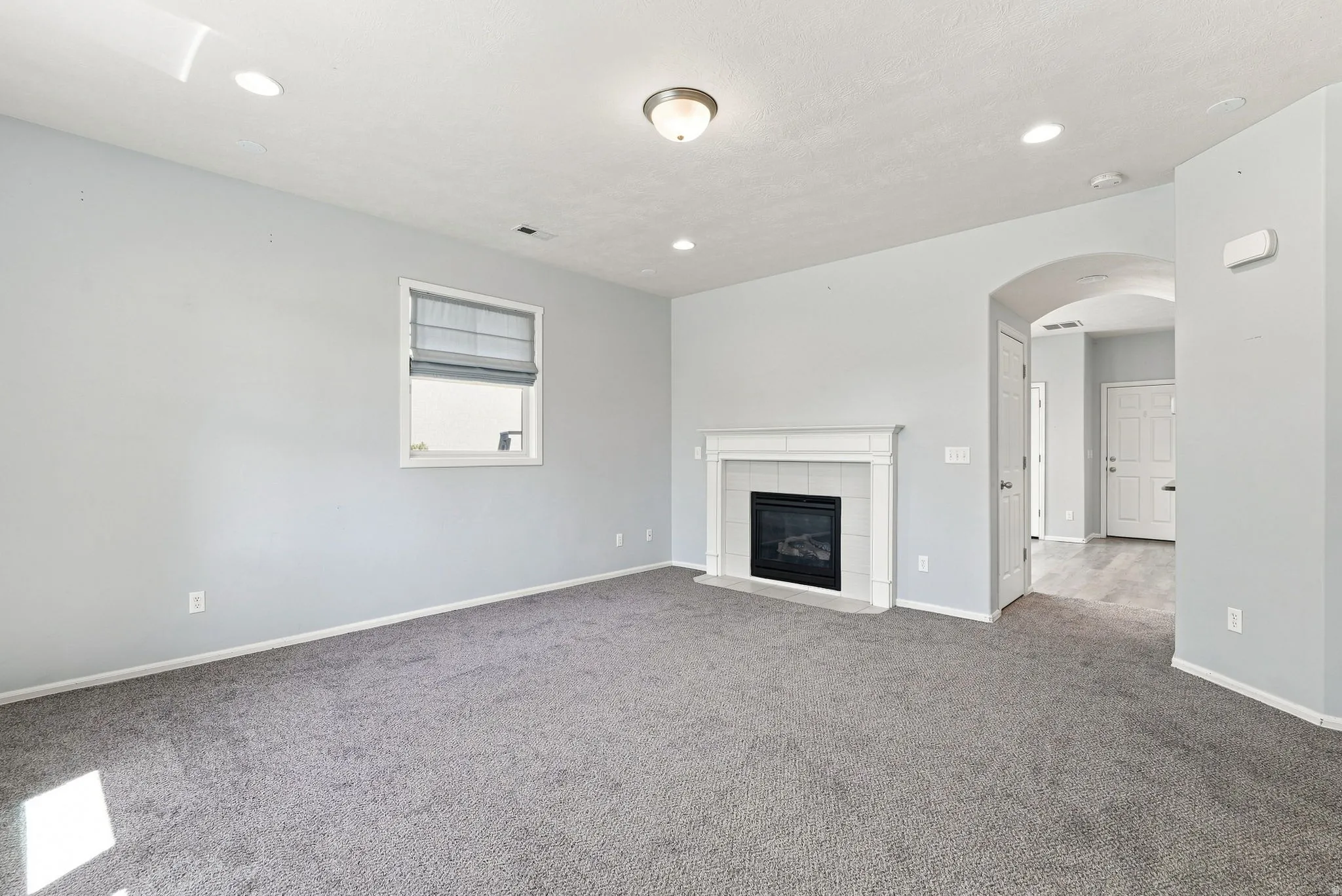 Wall-mounted window treatment, built-in fireplace with white mantel and dark tile surround, recessed lighting, ceiling-mounted light fixture, and archway with wood-finish flooring