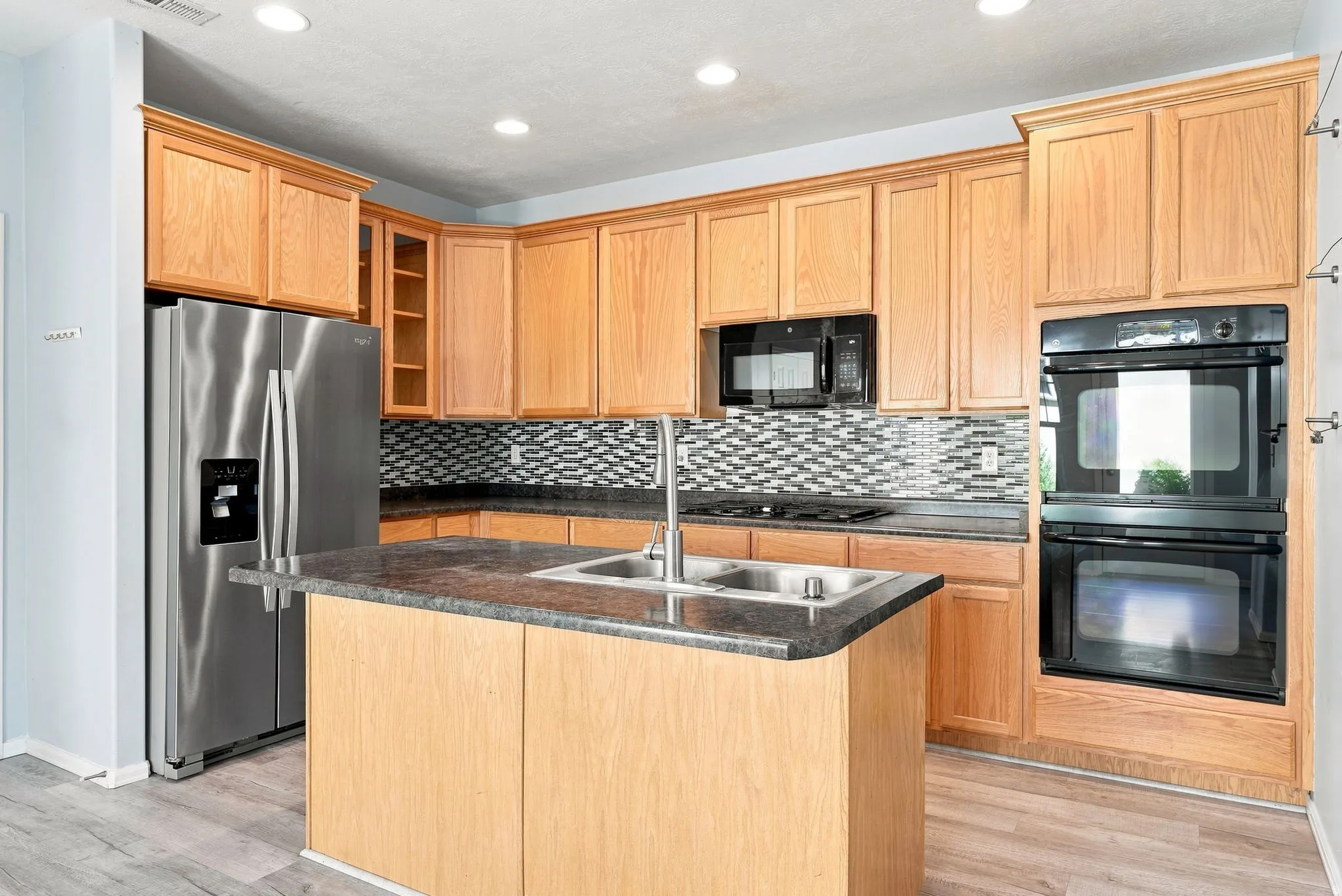Kitchen featuring wood cabinetry, stainless steel appliances, a center island with sink, black countertops, and a tiled backsplash