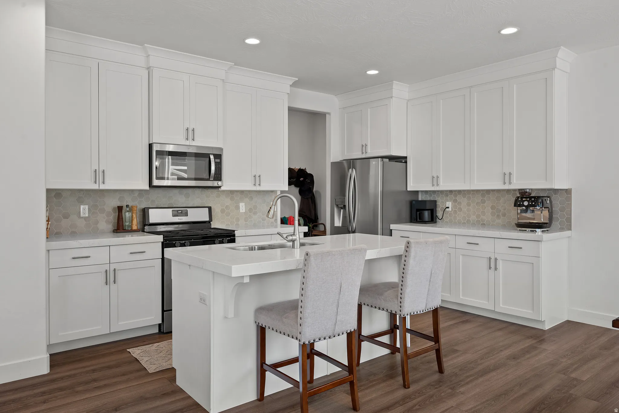 White shaker-style cabinetry with crown molding, stainless steel appliances, hexagon tile backsplash, center island with an undermount sink, and wood-finish flooring
