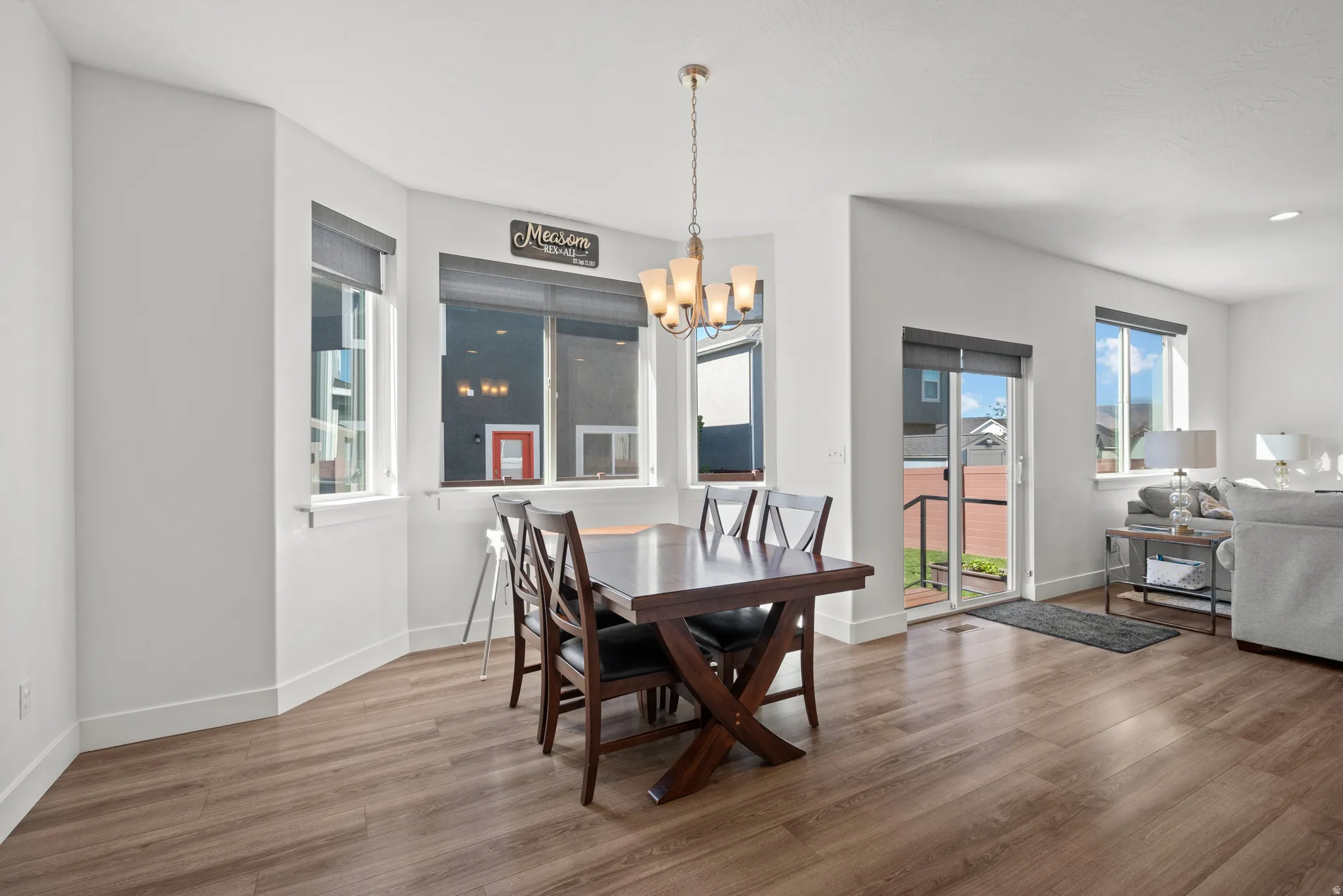 Wood-finish flooring throughout, bay window with multiple panes, central chandelier, sliding glass door, and white painted walls