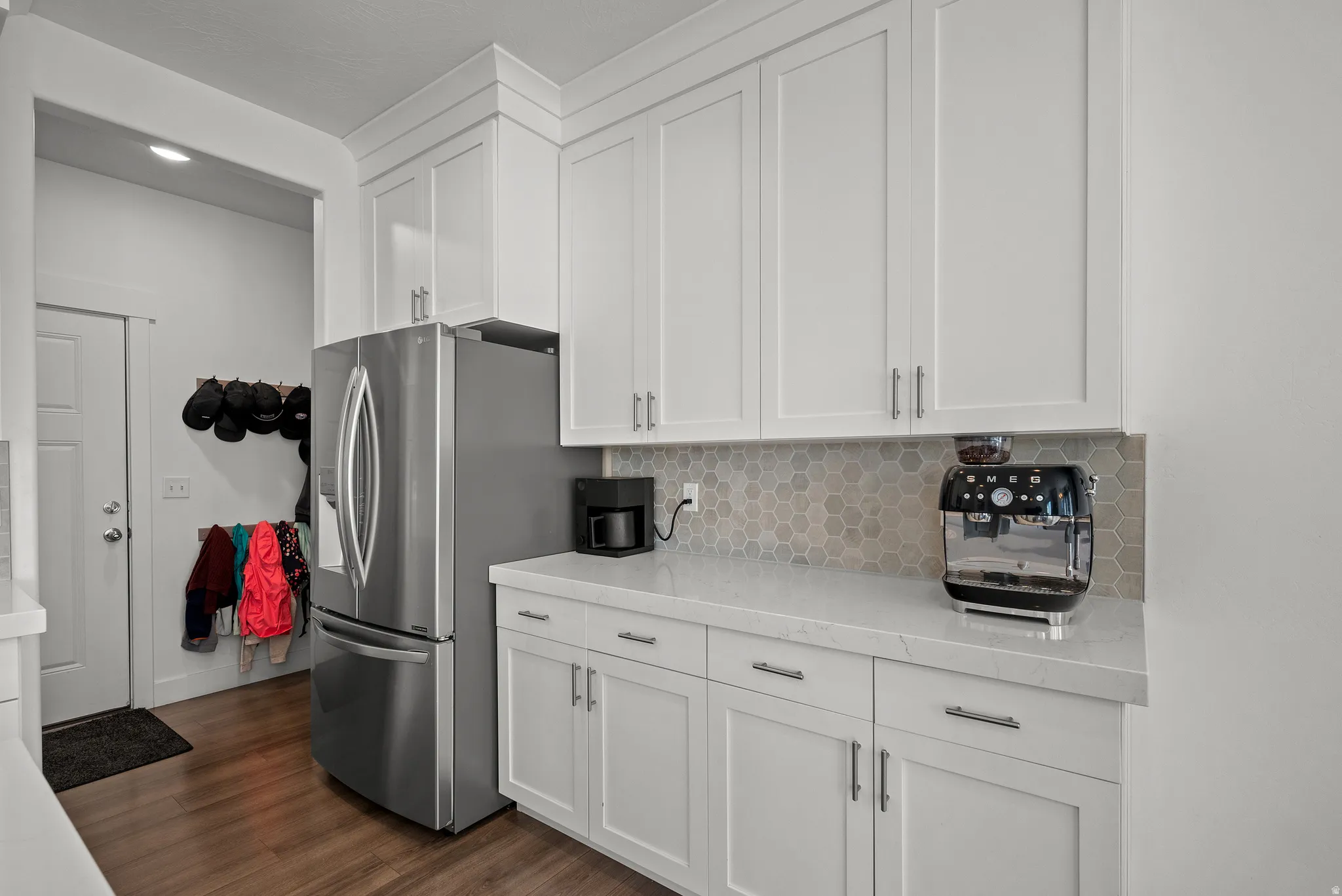 White shaker-style cabinetry, light-toned countertops, hexagonal tile backsplash, stainless steel refrigerator, and wood-finish flooring