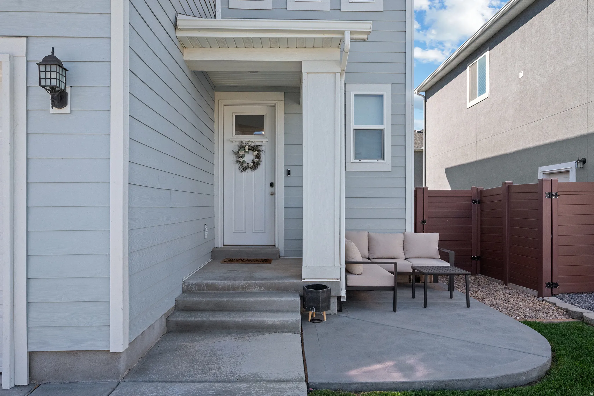 Light blue horizontal siding, white trim, covered porch entry, concrete patio, and brown privacy fence
