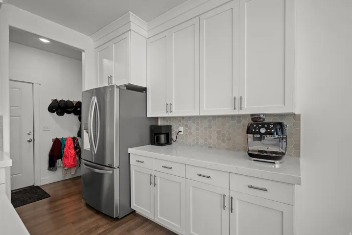 White shaker-style cabinetry, light-toned countertops, hexagonal tile backsplash, stainless steel refrigerator, and wood-finish flooring