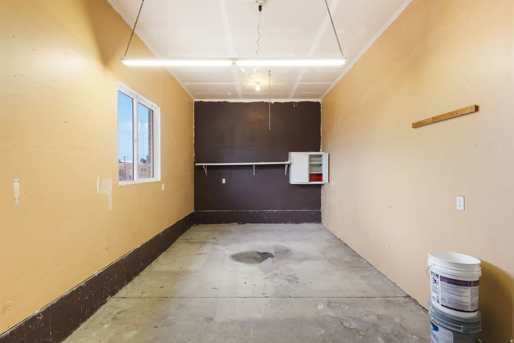 Concrete flooring, wall-mounted shelving, white framed window, permanent overhead lighting, and built-in wall cabinet