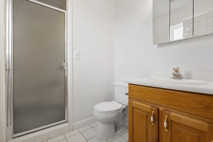 Shower with frosted glass enclosure, white tile flooring, wood-finish vanity with countertop, and wall-mounted mirrored medicine cabinet