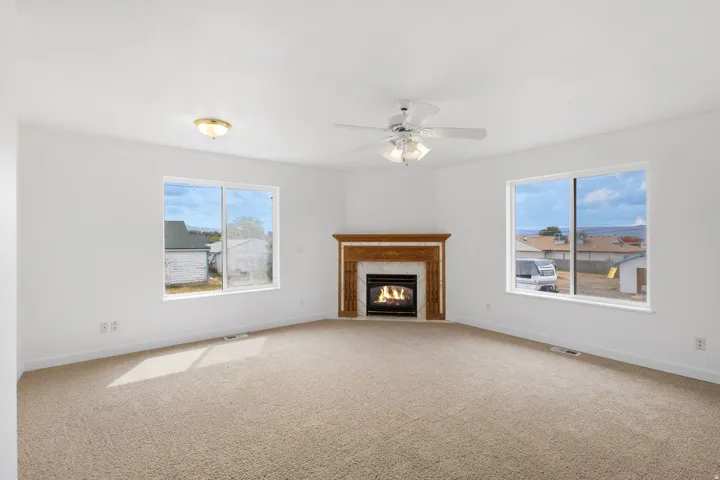 Carpeted living area featuring a fireplace with a wood-finish mantel and tile surround, a ceiling fan with integrated lighting, and two large windows