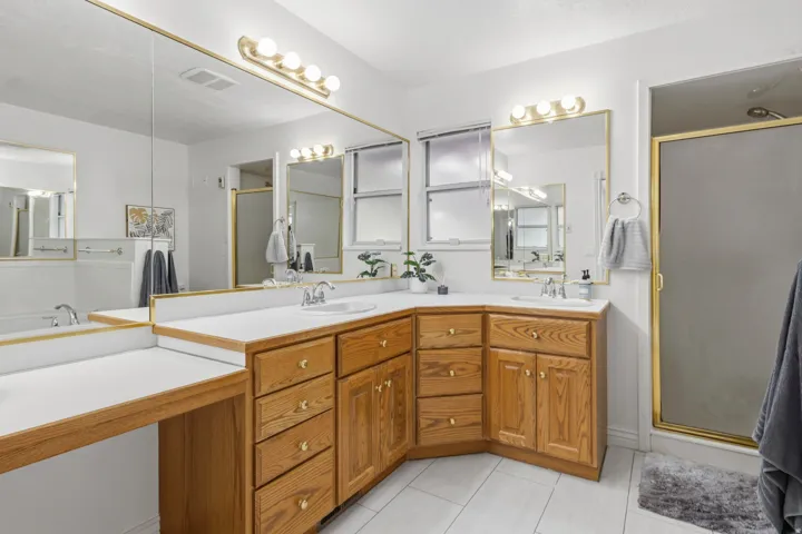 Dual vanity with light wood cabinetry and white countertops, large wall-mounted mirrors, and an enclosed shower with a gold-toned frame