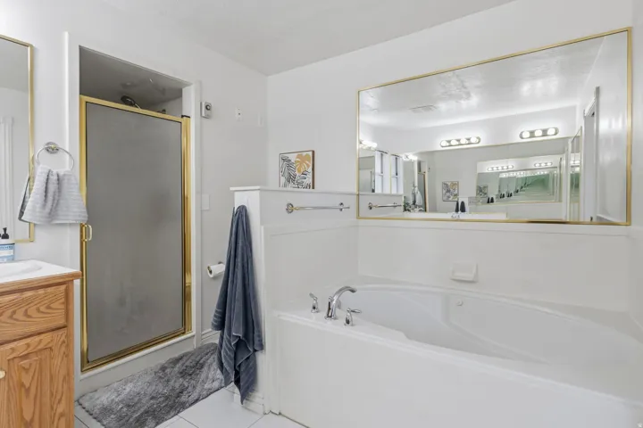 Built-in soaking tub, large vanity mirror, wood-finish cabinetry, white tile flooring, and a separate shower with a brass-finish frame