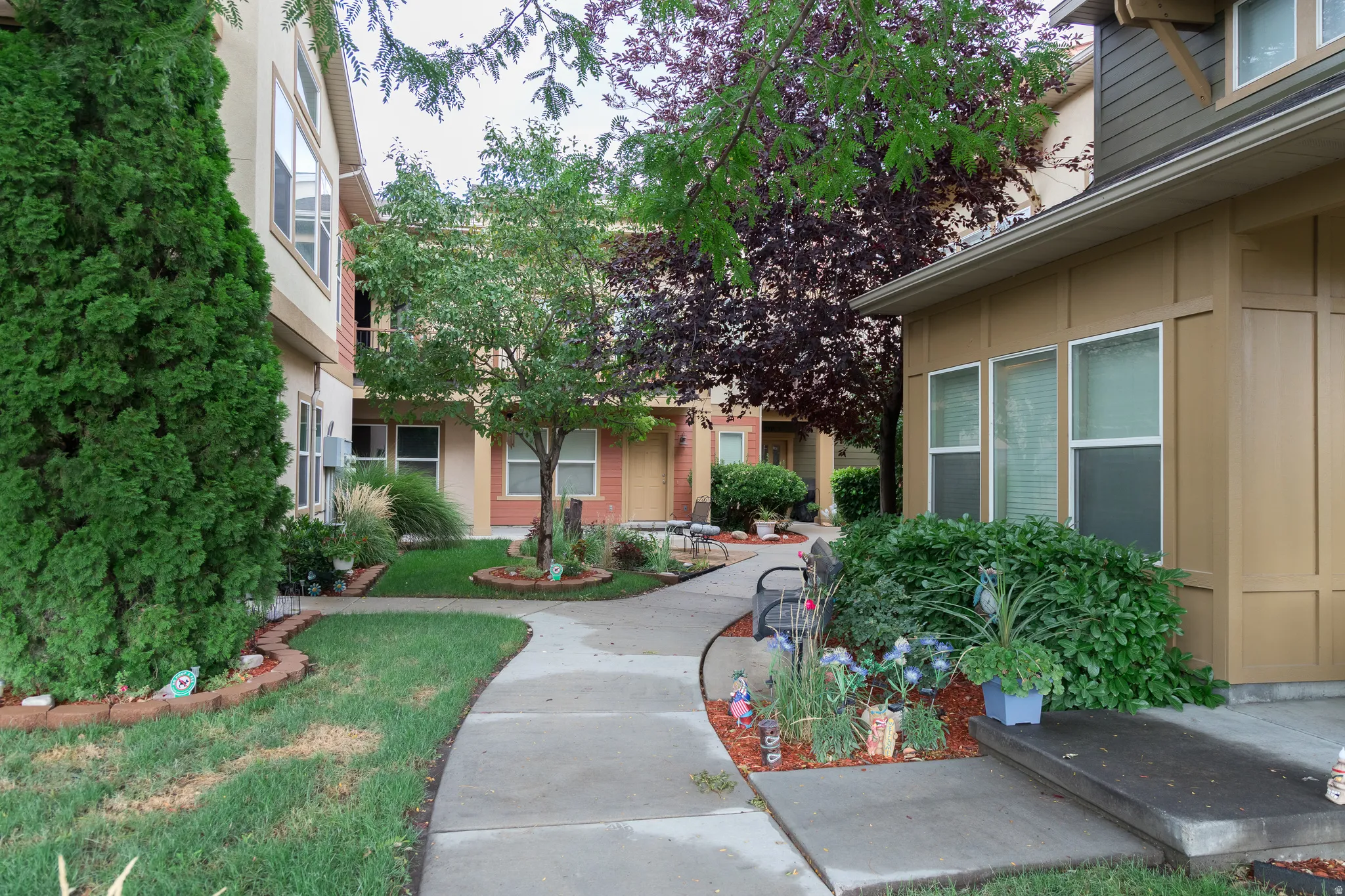 Exterior featuring multiple windows, a concrete pathway, landscaped garden beds, and mature trees