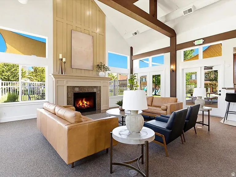Vaulted ceiling with exposed wood-finish beams, fireplace with stone surround, extensive window placement, board and batten accent wall, and double glass doors