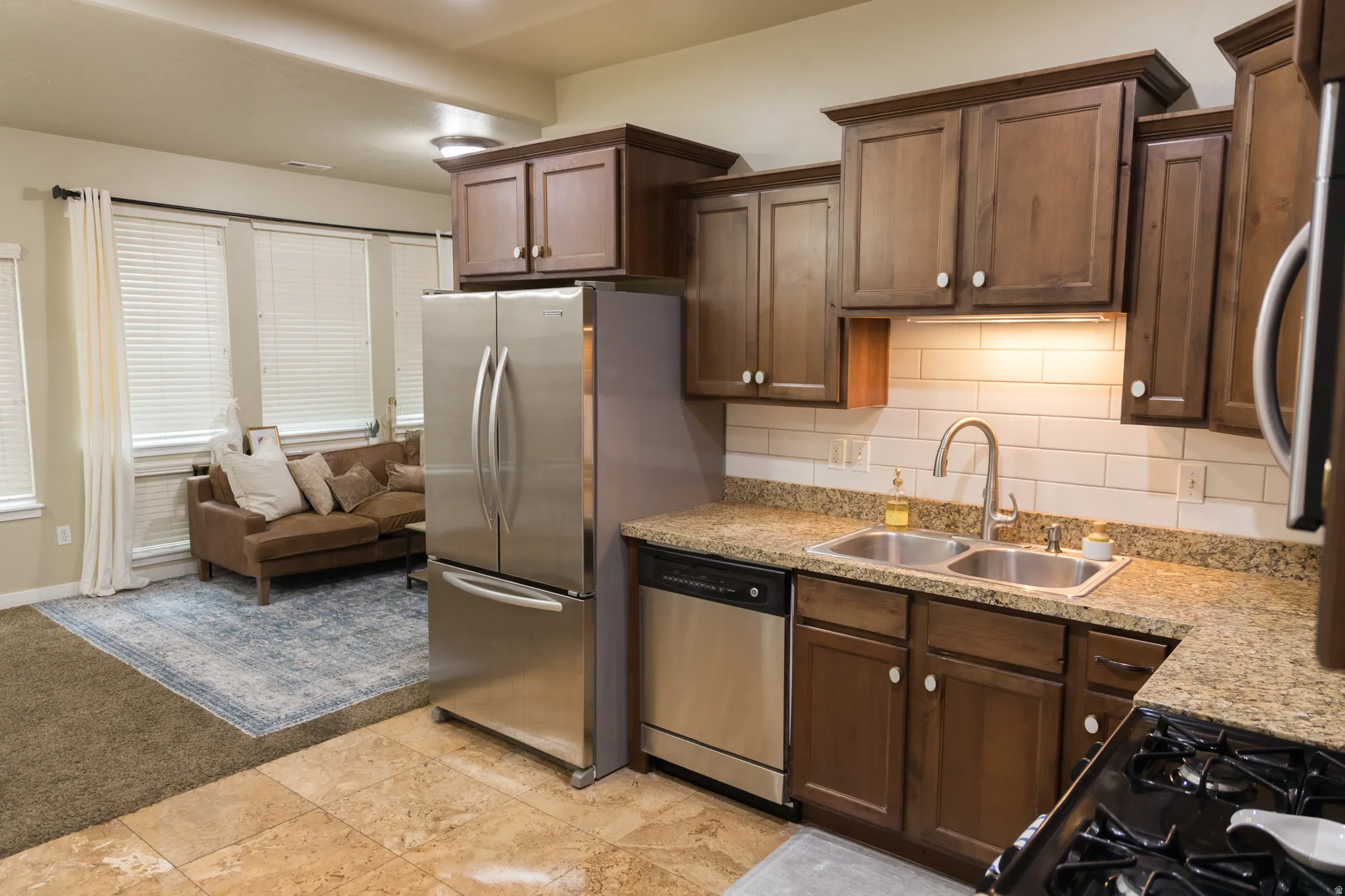 Kitchen with wood-finish cabinetry, stone countertops, dual stainless steel sink, and subway tile backsplash