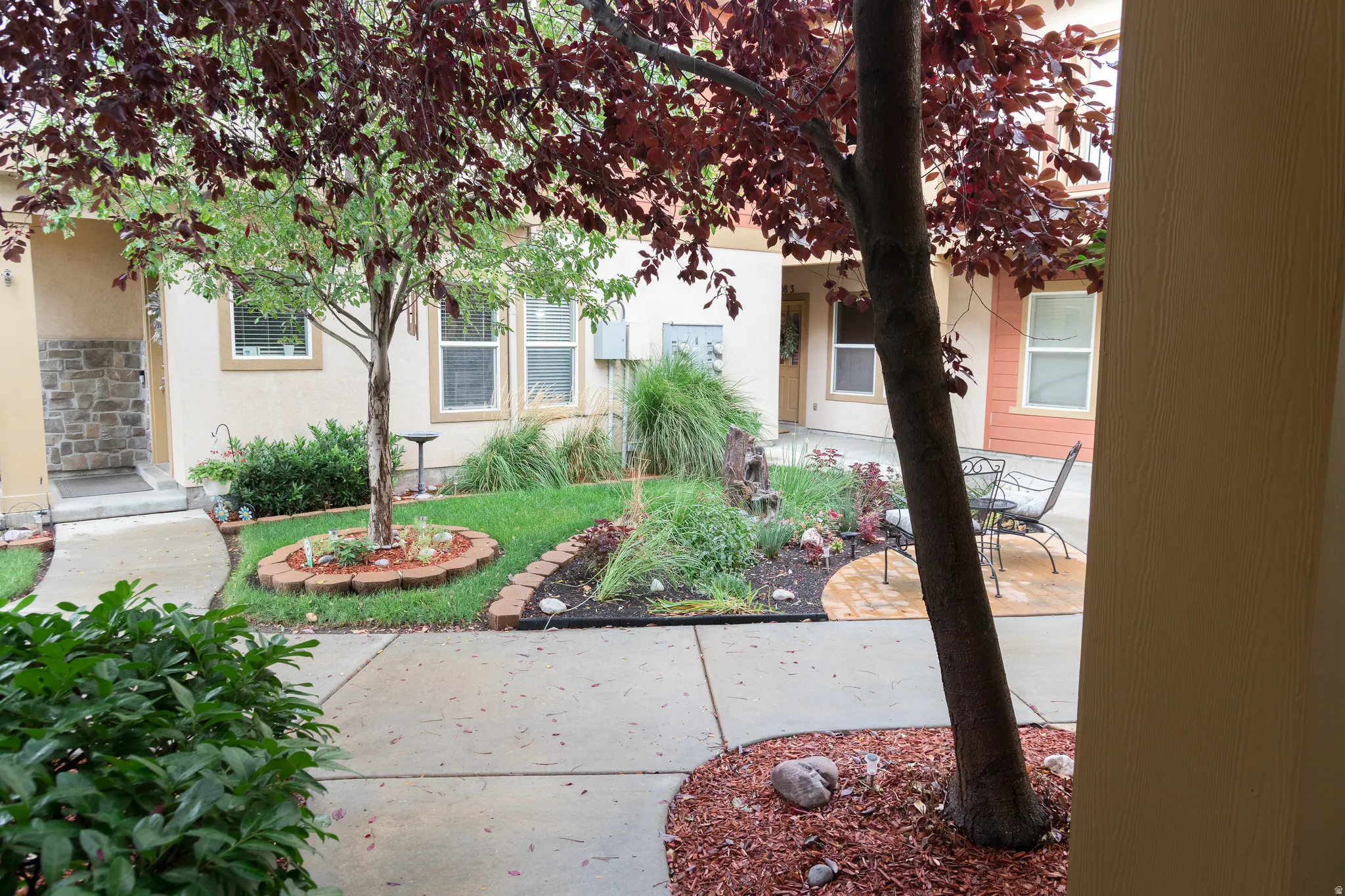 Courtyard entrance featuring a paved walkway, mature trees, landscaped garden beds, and a stone facade accent