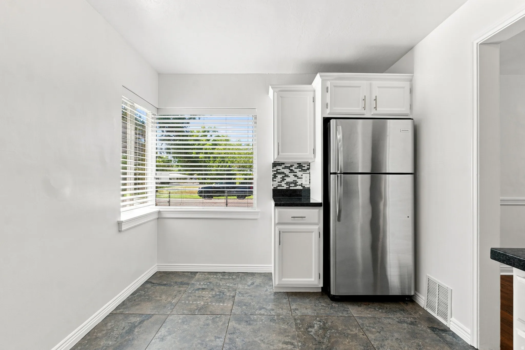 Stainless steel refrigerator, white cabinetry, dark countertop, mosaic tile backsplash, and large window with blinds