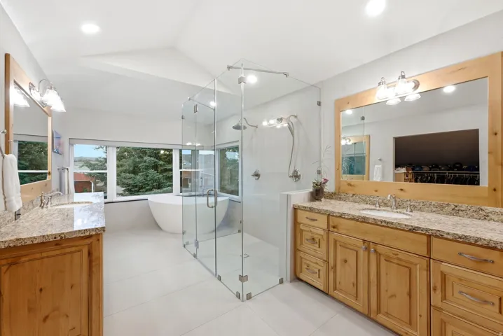 Dual wood-finish vanities with stone countertops, a large glass-enclosed shower, a freestanding soaking tub, and multiple windows