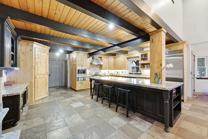 Kitchen featuring an expansive island with seating, natural wood cabinetry, stainless steel appliances, exposed wood beams, and a tile floor