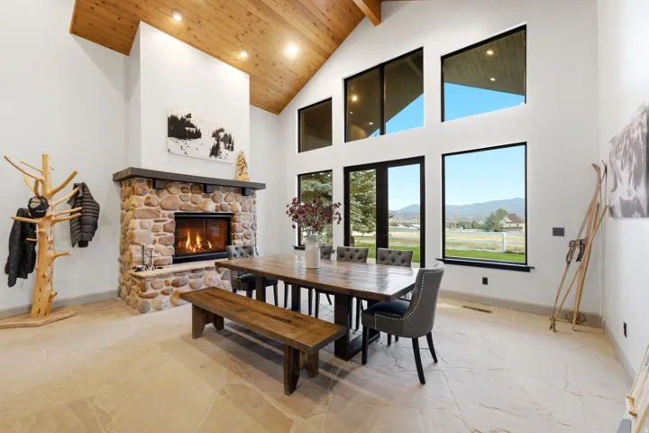 Vaulted wood-finish ceiling, stone fireplace with hearth, multiple large windows, and light-toned tile flooring