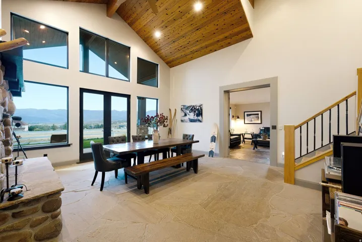 Vaulted wood-paneled ceiling with exposed beams, stone-clad fireplace with mantel, large windows and glass French doors, natural stone flooring, and a staircase with wood newel posts and wrought iron balusters