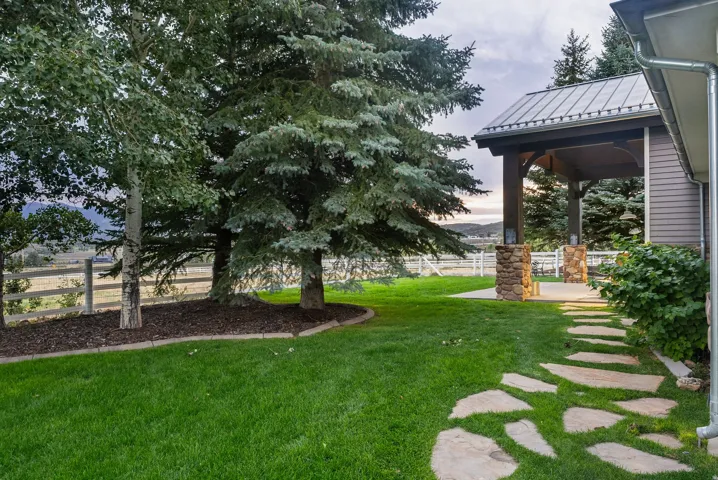 Lush green lawn with mature trees and a flagstone pathway leading to a covered patio featuring stone-clad columns and a metal roof